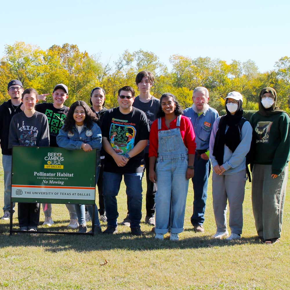A group of students standing in a field next to sign which reads “Bee Campus USA. UT Dallas. Pollinator Habitat. No Mowing.”.