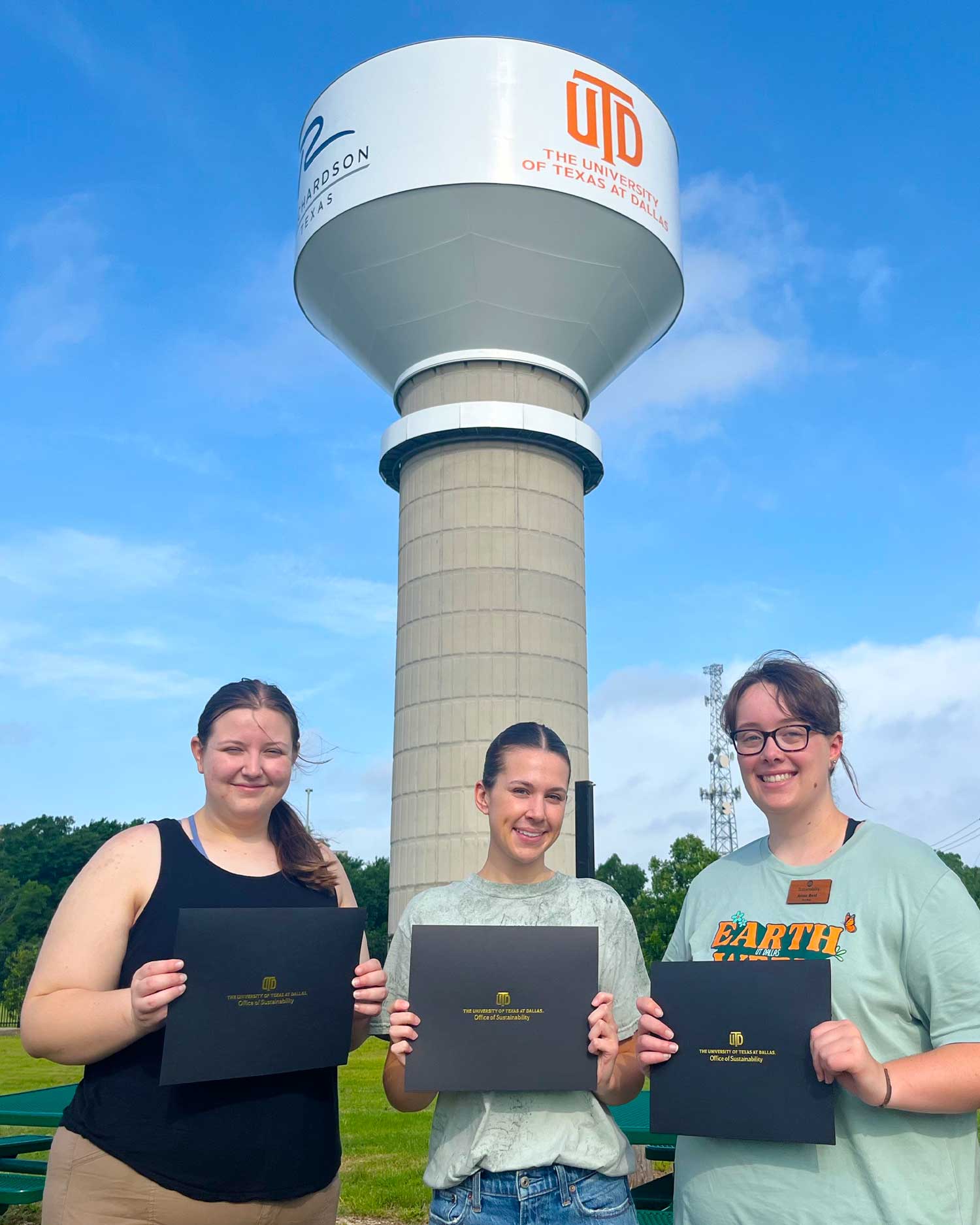 Three students stand under the water tower at the UTD Eco Hub, holding Sustainability Honors certificates.