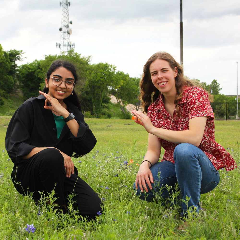 Two people making the UT Dallas “Mini Whoosh” symbol with their left hands, kneeling in a field of wildflowers.
