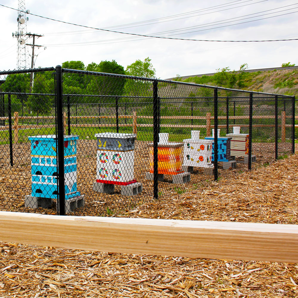 A row of tall wooden boxes standing on brick feet, each one painted in a different colorful design. The boxes are inside a high black chain-link fence, which is itself inside a shorter wooden fence.