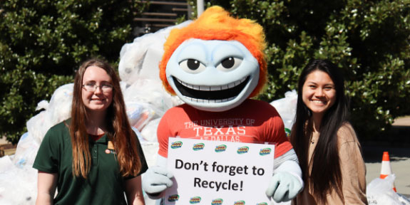 Campus Race to Zero Waste. School mascot Temoc and two students stand in front of a pile of filled recycling bags holding up a sign which reads “Don’t forget to recycle!”