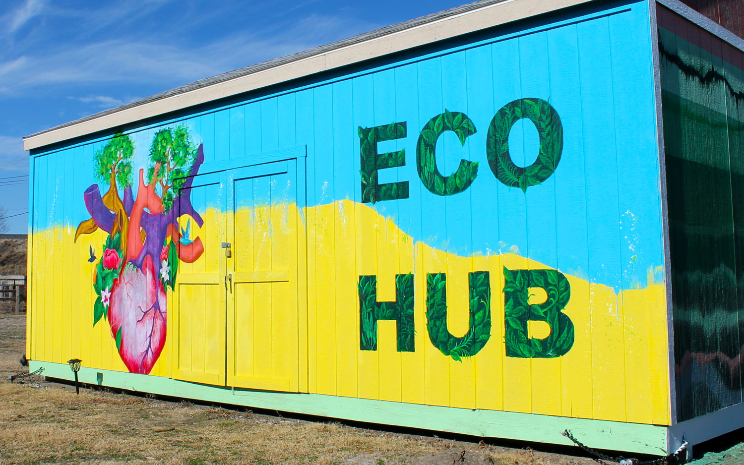 The side of a single-story gabled-roof building. The top half of the background is painted light blue, the bottom half is painted yellow, with a blurred boundry between them. To the left is a human heart, from which sprout trees and flowers that have attracted humingbirds. To the right are the words “ECO HUB” in letters made of greenery.