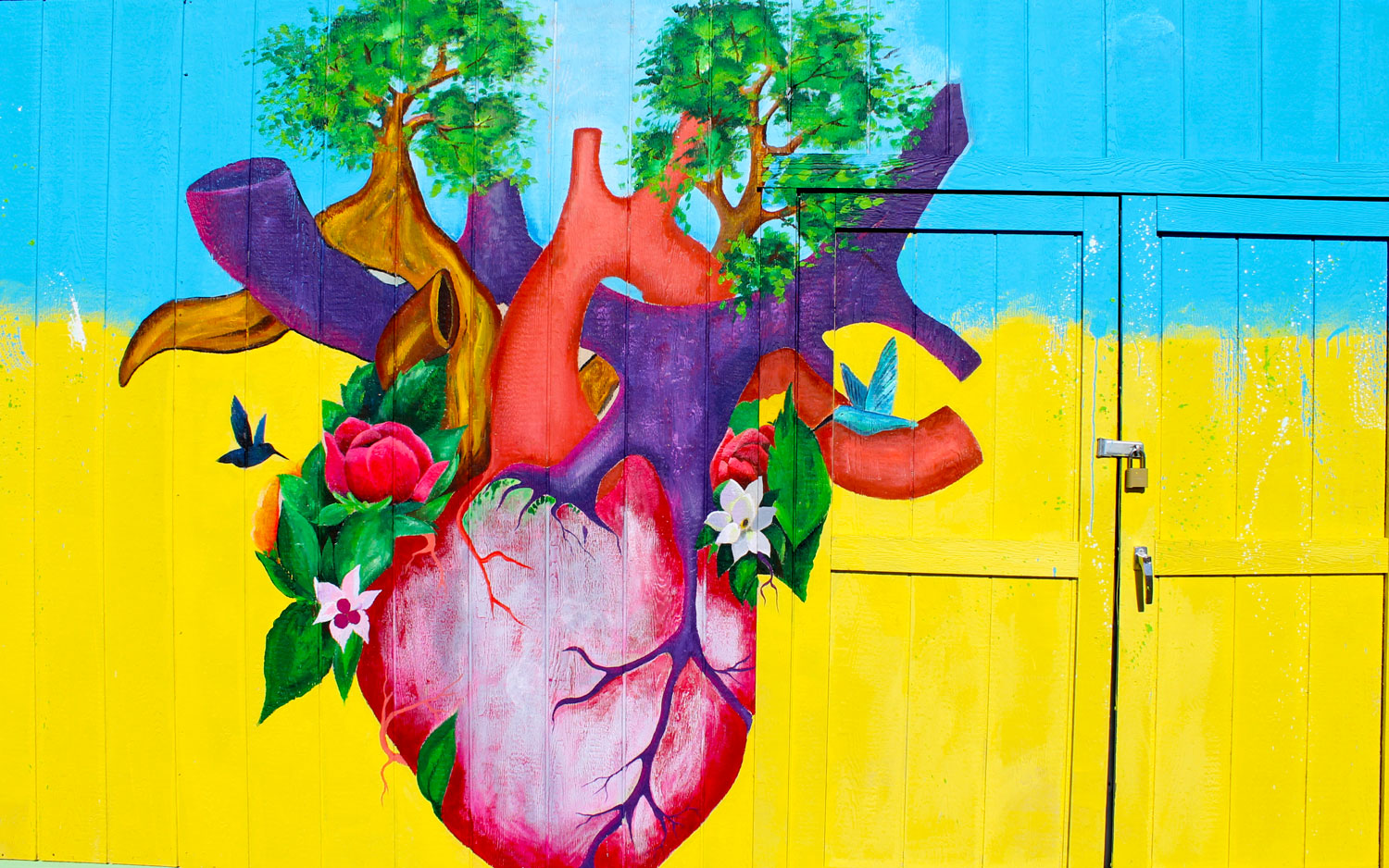 Close-up view of a building wall and double doors. The top half of the background is painted light blue, the bottom half is painted yellow, with a blurred boundry between them. In the center is a human heart, from which sprout trees and flowers that have attracted humingbirds.