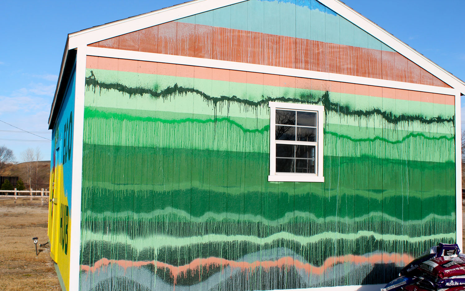 The side of a single-story gabled-roof building, painted in wide horizontal stripes overlapped by jagged dripping sprayed-on lines, in shades of green, blue, and terra cotta.