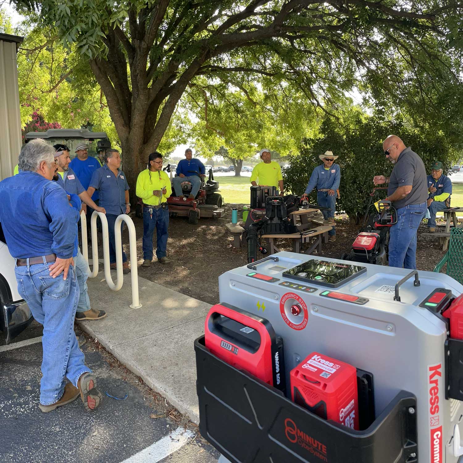 A group of men gather under a tree to examine a variety of electric gardening tools laid out across a table.