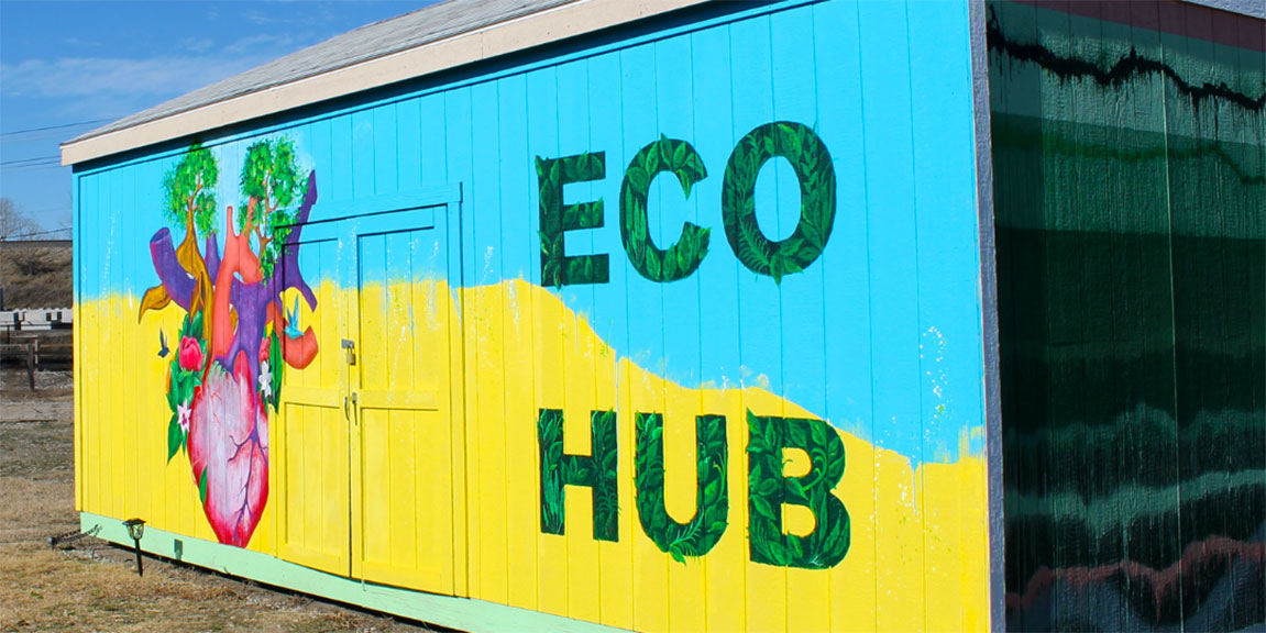 The side of a single-story gabled-roof building. The top half of the background is painted light blue, the bottom half is painted yellow, with a blurred boundry between them. To the left is a human heart, from which sprout trees and flowers that have attracted humingbirds. To the right are the words “ECO HUB” in letters made of greenery.