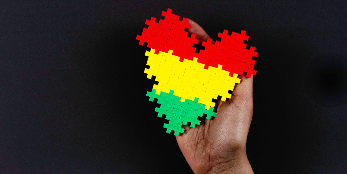 A hand reaching out of the darkness, holding a heart-shaped section of jigsaw puzzle stiped with the colors of Black Liberation.