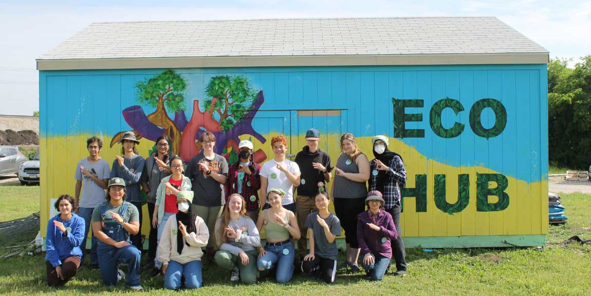 A group of students standing in front of the colorful Eco Hub, shed making the sign of the UTD mini-whoosh with their left hands.