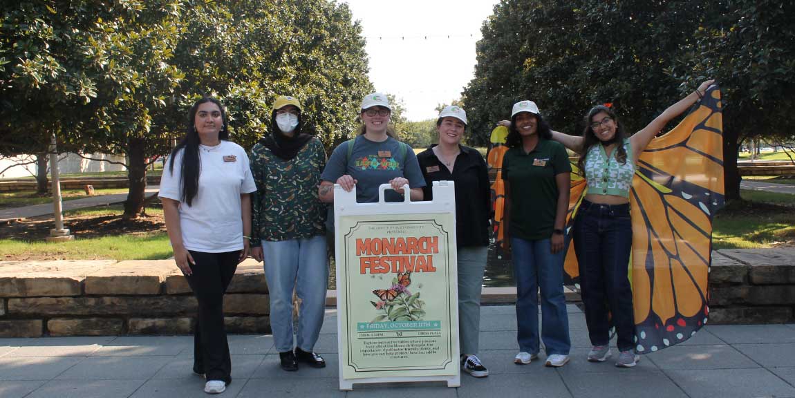 A group of students standing behind a sign reading “Monarch Festival”. On wears a pair on monarch butterfly wings.