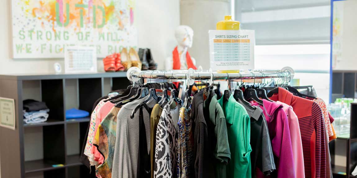 A room with clothes and shoes laid out on shelves along its walls, a circular clothes rack full of clothes on hangers in its center, a manequin modeling a scarf in the back, and size charts posted throughout the space.