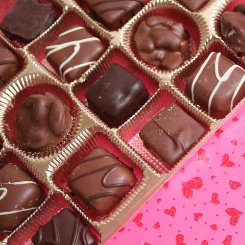 An open gift box of chocolates on a pink background sprinkled with red hearts.