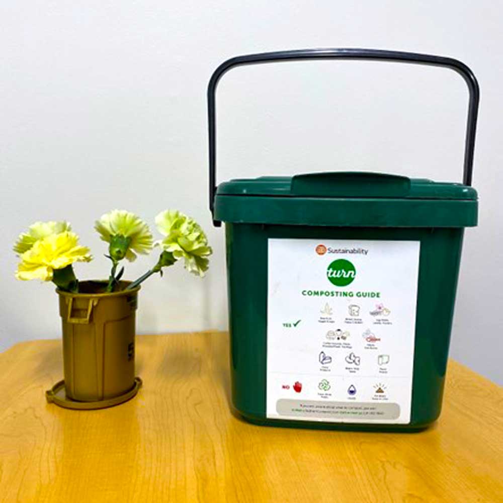 A small UT Dallas composting bin bearing a label with a composting guide, sitting on a table next to a bouquet of flowers.