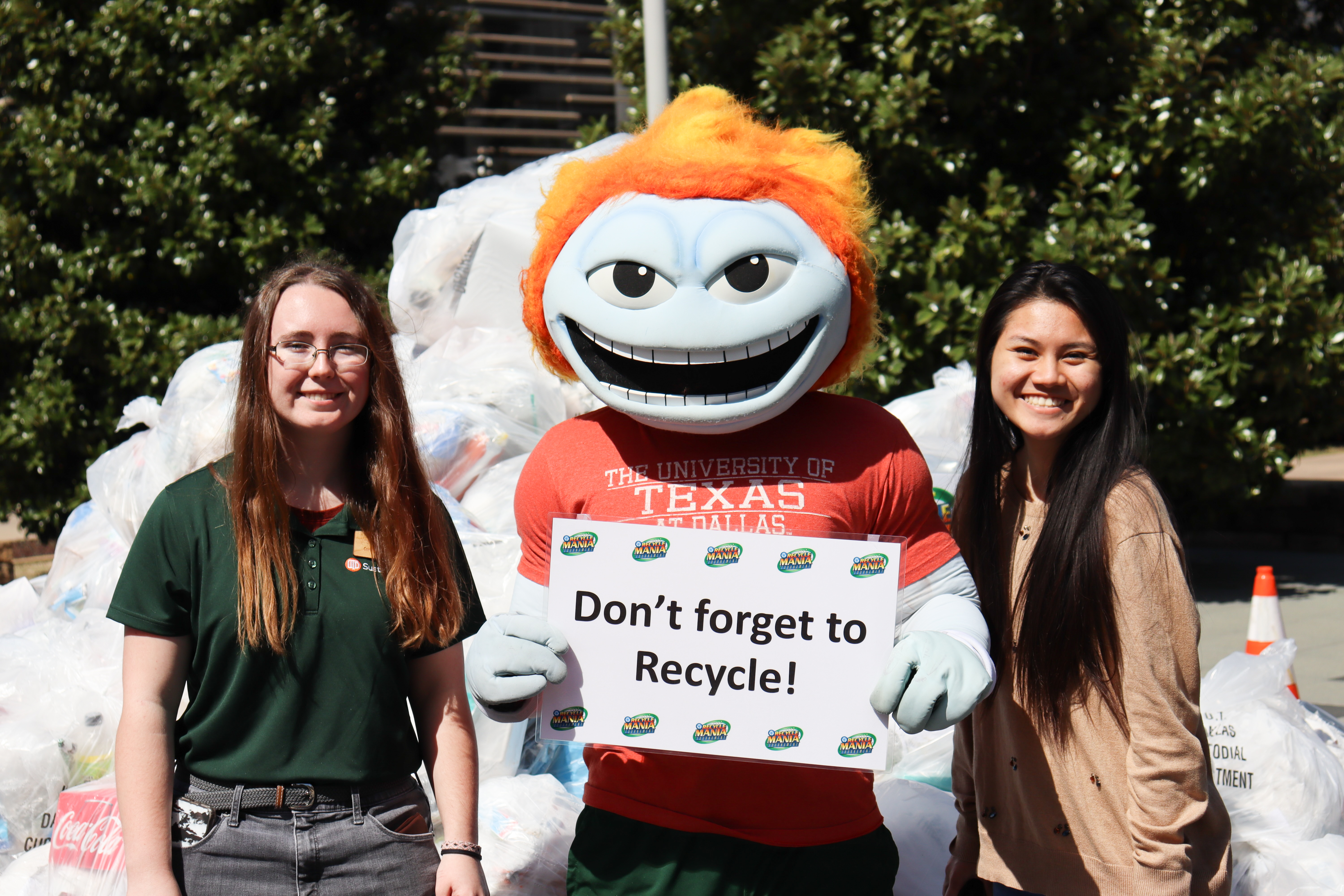 Two students and Temoc, the University mascot, stand in front of a pile of recyclables while Temoc holds a sign the reads “Don't Forget to Recycle”