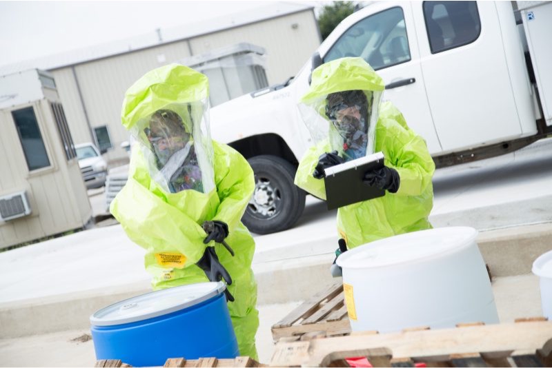 Two workers in HAZMAT suits inspect barrels of chemicals.