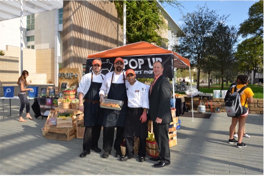 Three men in kitchen uniforms and one man in a suit in front of a stand full of baskets of food.