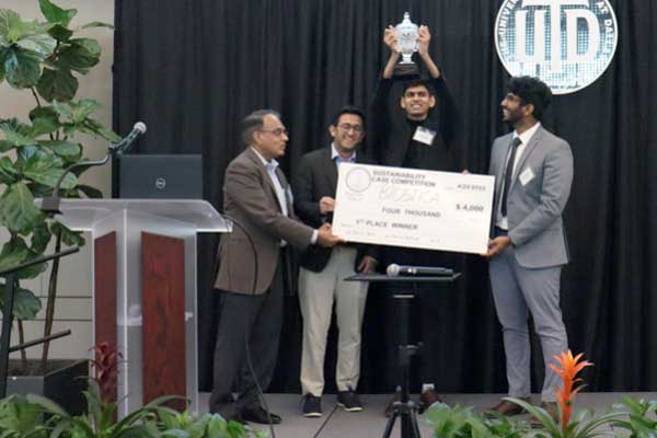 A man on a stage hands a large check to two students while a third student raises a trophy behind them.